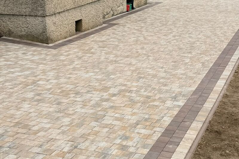 Podjazd z kostki brukowej w kolorze piaskowym z brązowym pasem obramowania, układany wzdłuż ściany budynku